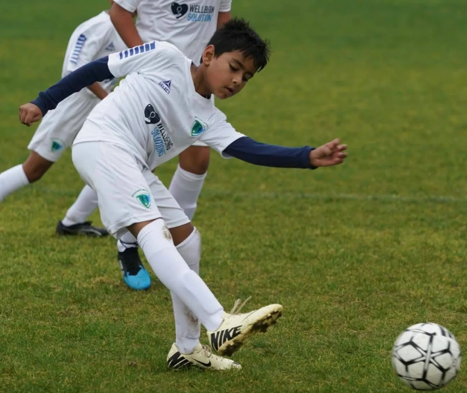Young footballer kicking the ball in a white kit bearing the WellbeingSolutions logo