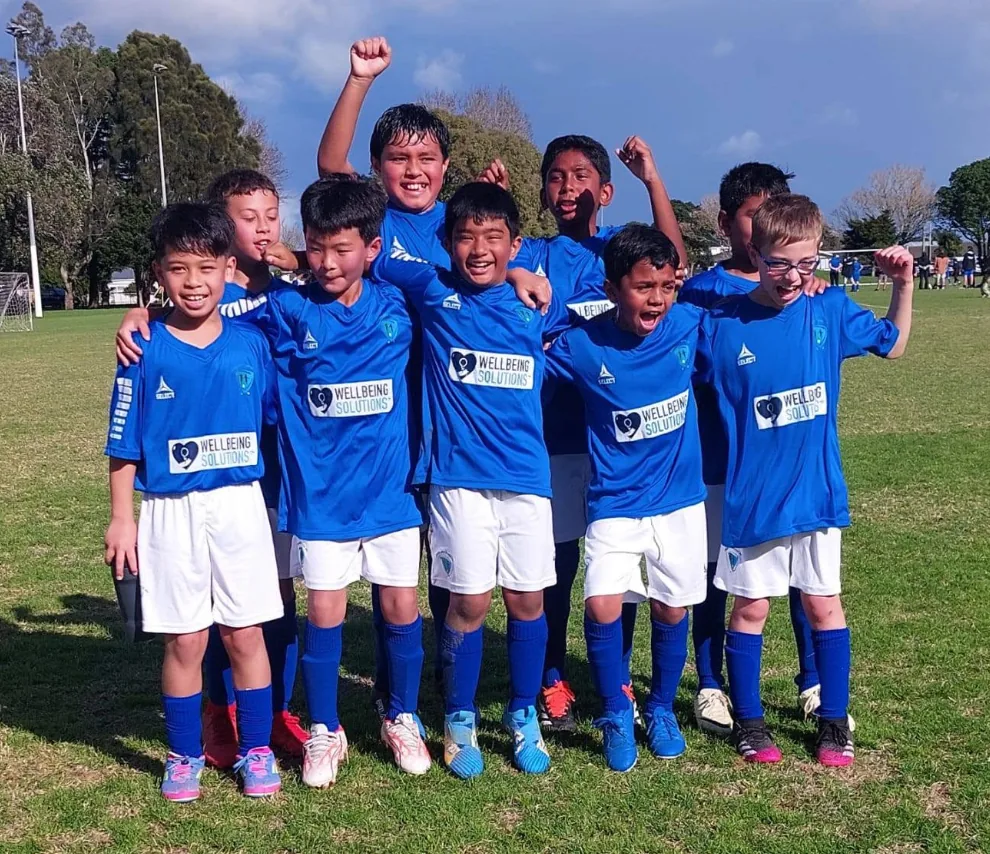 Children celebrating together in blue football shirts displaying the WellbeingSolutions sponsor logo